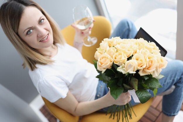 Jeune femme avec bouquet de roses thé assis à l'ordinateur portable avec un verre de vin blanc