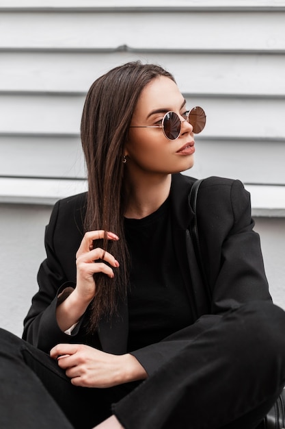 Jeune femme branchée hipster dans des lunettes de soleil élégantes dans des vêtements noirs pour jeunes à la mode s'asseoir près d'un mur vintage gris en bois à l'extérieur. Le mannequin jolie fille magnifique se détend dans la rue.