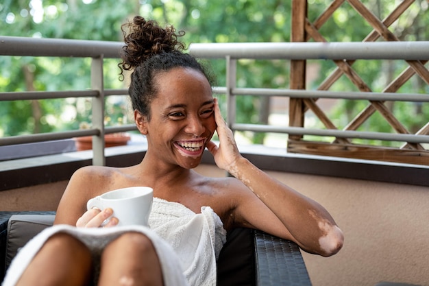 Jeune femme brésilienne africaine souriant et buvant un verre pendant un moment d'insouciance au spa femme avec vitiligo se détendre en vacances sur le balcon après la douche