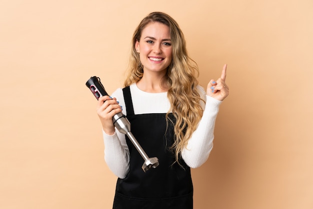 Jeune femme brésilienne à l'aide d'un mixeur plongeant isolé sur beige pointant vers le haut une excellente idée