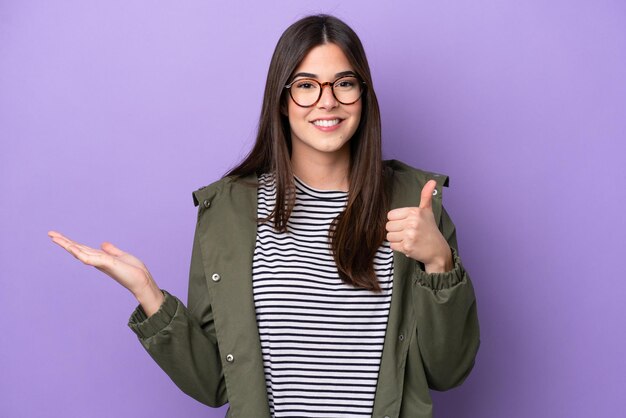 Jeune femme brésilienne isolée sur fond violet tenant un fond imaginaire sur la paume pour insérer une annonce et avec les pouces vers le haut