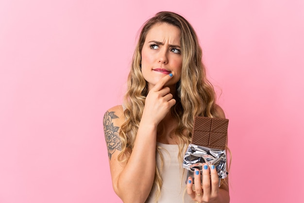 Jeune femme brésilienne isolée sur rose en prenant une tablette de chocolat et ayant des doutes