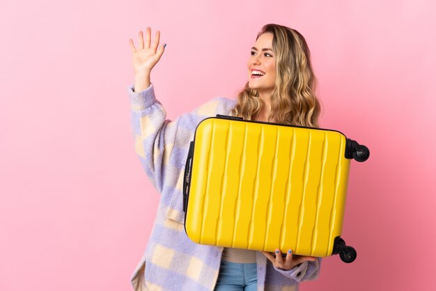 Jeune femme brésilienne isolée sur rose en vacances avec valise de voyage et saluant