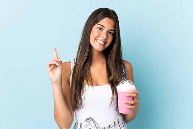 Jeune femme brésilienne avec milk-shake aux fraises isolé sur mur bleu montrant et en soulevant un doigt en signe de la meilleure