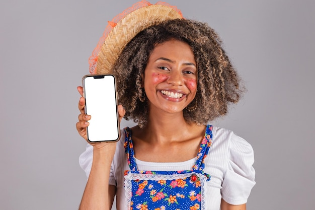 Jeune femme brésilienne noire avec des vêtements de campagne habillés pour Festa Junina tenant un smartphone avec écran blanc pour les applications mobiles publicitaires