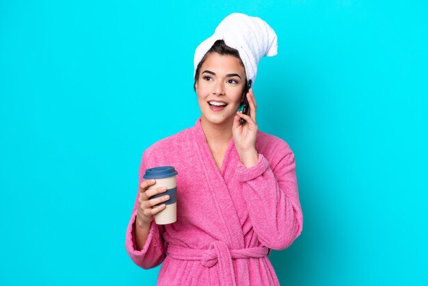 Jeune femme brésilienne avec un peignoir isolé sur fond bleu tenant du café à emporter et un mobile