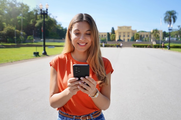 Jeune femme brésilienne riant et envoyant des SMS sur son téléphone portable marchant à l'extérieur par une journée ensoleillée