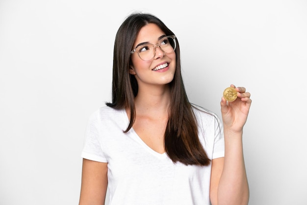 Jeune femme brésilienne tenant un bitcoin isolé sur fond blanc levant les yeux en souriant