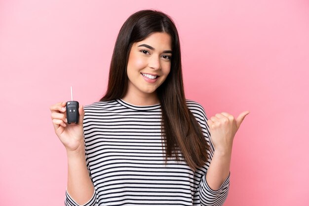 Jeune femme brésilienne tenant des clés de voiture isolées sur fond rose pointant vers le côté pour présenter un produit