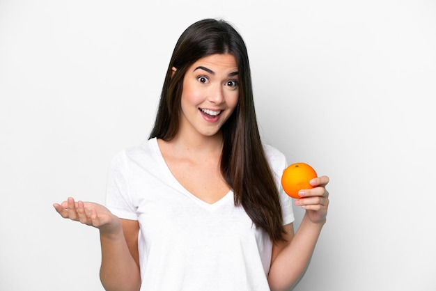 Jeune femme brésilienne tenant une orange isolée sur fond blanc avec une expression faciale choquée