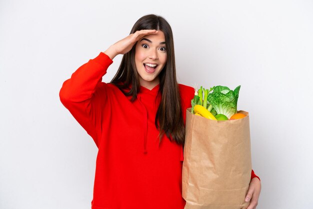 Jeune femme brésilienne tenant un sac d'épicerie isolé sur fond blanc avec une expression de surprise