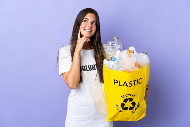 Jeune femme brésilienne tenant un sac plein de bouteilles en plastique à recycler isolé sur mur violet en pensant à une idée tout en levant les yeux