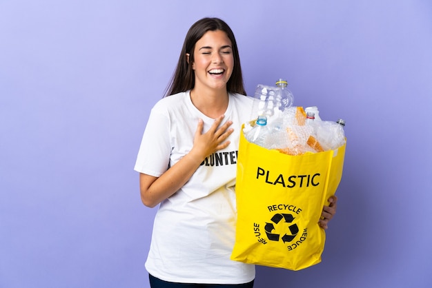 Jeune femme brésilienne tenant un sac plein de bouteilles en plastique à recycler isolé sur violet souriant beaucoup