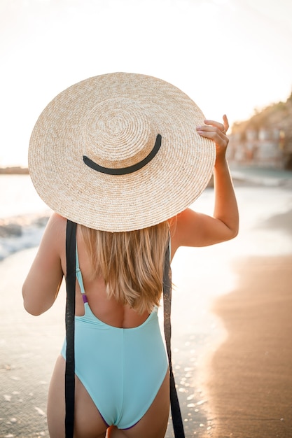 Une jeune femme bronzée dans un beau maillot de bain avec un chapeau de paille se dresse et se repose sur une plage tropicale de sable et regarde le coucher de soleil et la mer. Mise au point sélective. Concept de vacances au bord de la mer