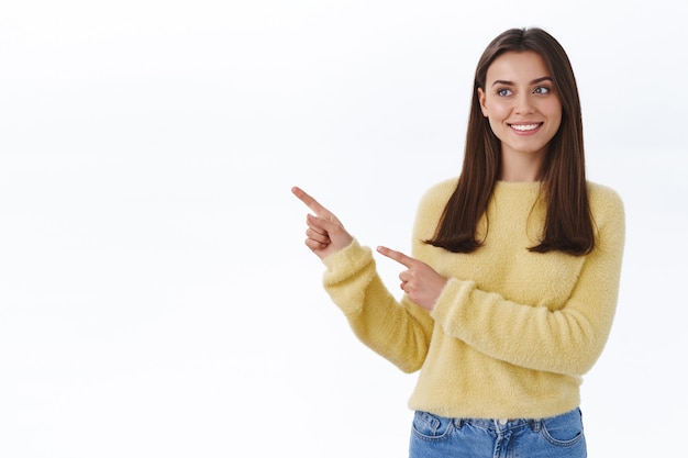 Une jeune femme brune adulte satisfaite fait la promotion du produit, pointant et regardant vers la gauche avec un sourire heureux et ravi comme donner une recommandation où trouver tout ce dont vous avez besoin, un mur blanc debout satisfait