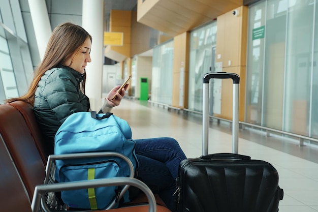 Jeune femme brune à l'aéroport attend son vol quelque chose regardant smartphone Elle est assise à côté de la vue latérale de la valise et du sac à dos