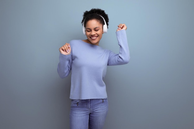 Jeune femme brune afro énergique écoutant de la musique à l'aide d'un casque sans fil blanc en studio