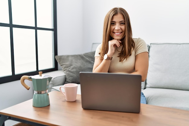 Jeune femme brune à l'aide d'un ordinateur portable à la maison en buvant une tasse de café en regardant la caméra en souriant avec les bras croisés et la main levée sur le menton en pensant positif