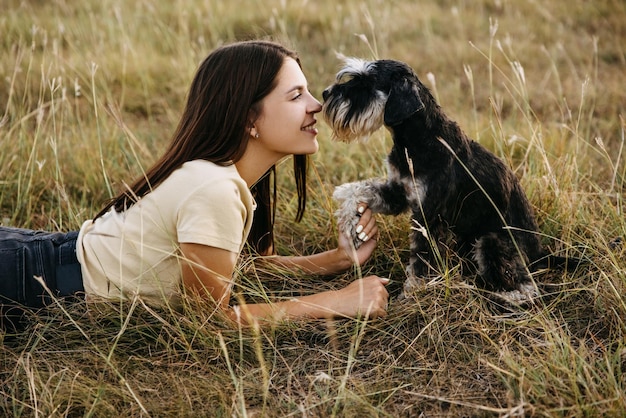 Une jeune femme brune allongée dans un champ avec un chien de race schnauzer en miniature touchant le nez