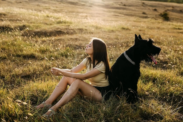 Une jeune femme brune assise dans un champ à côté d'un grand chien noir de race géante schnauzer