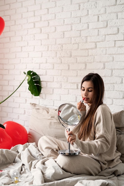 Jeune femme brune assise dans le lit en train de colorer les lèvres