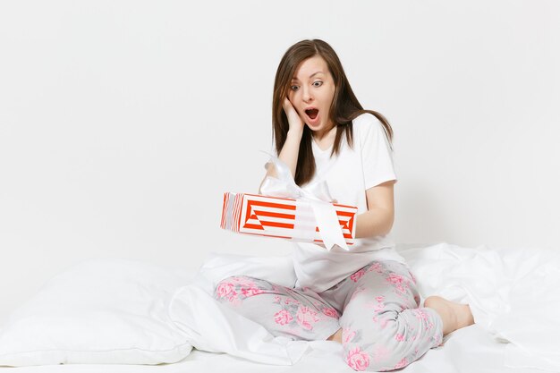 Jeune femme brune assise dans son lit avec boîte-cadeau à rayures rouges, drap blanc, oreiller, emballage dans une couverture isolée sur mur blanc