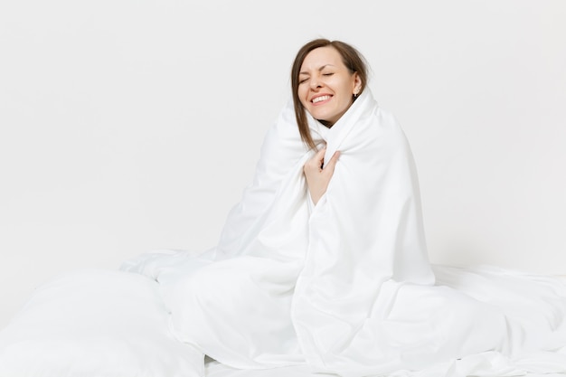 Jeune femme brune assise dans son lit avec un drap blanc, un oreiller, s'enveloppant dans une couverture sur un mur blanc