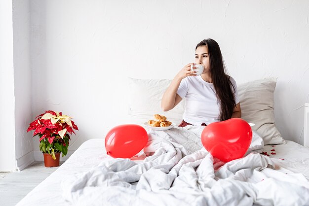 Jeune femme brune assise éveillée dans le lit avec des ballons en forme de coeur rouge et des décorations de boire du café manger des croissants