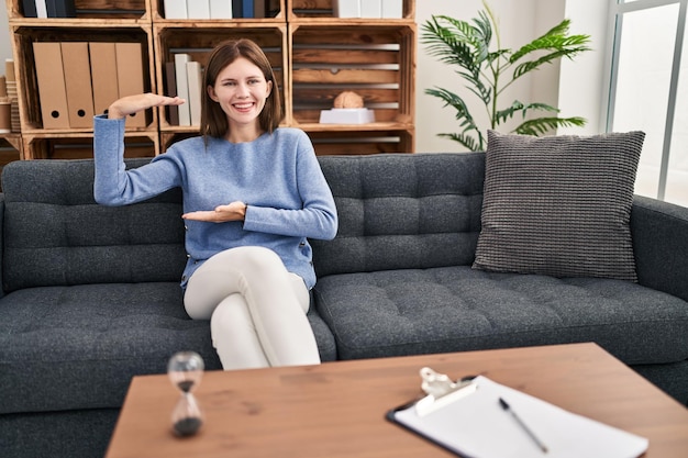 Jeune femme brune au bureau de consultation gesticulant avec les mains montrant le symbole de mesure de signe de grande et grande taille souriant en regardant le concept de mesure de la caméra