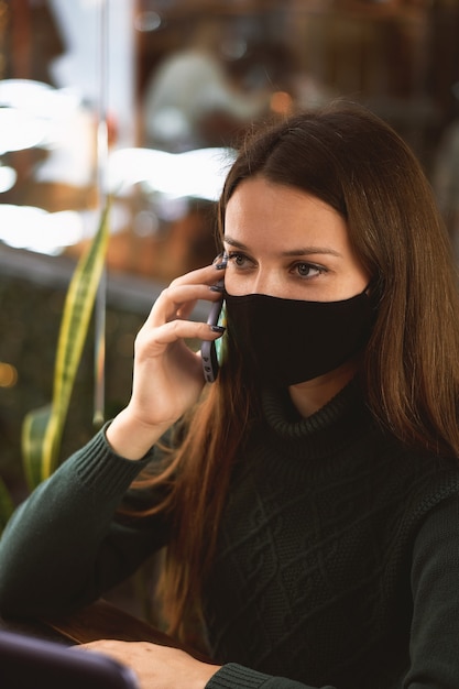 Jeune femme brune au masque médical au café avec ordinateur portable et smartphone