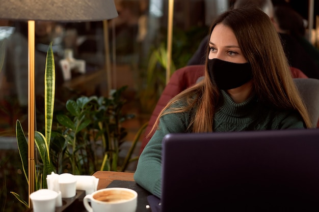 Jeune femme brune au masque médical au café avec ordinateur portable