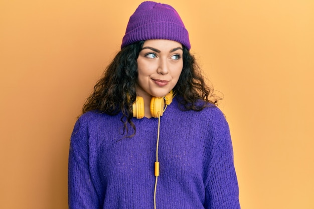 Jeune femme brune aux cheveux bouclés écoutant de la musique à l'aide d'un casque souriant regardant sur le côté et regardant loin en pensant