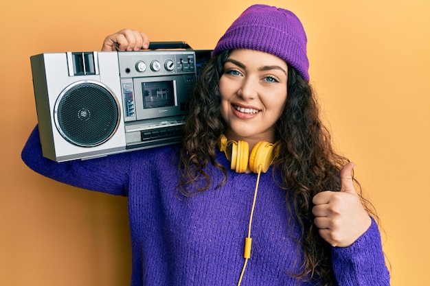 Jeune femme brune aux cheveux bouclés tenant une boombox écoutant de la musique souriant heureux et positif pouce vers le haut faisant un excellent signe d'approbation