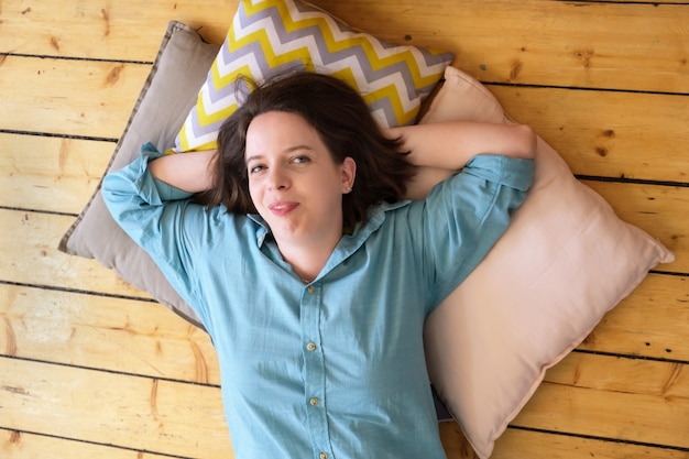 Jeune femme brune aux cheveux mi-longs est allongée sur des oreillers, la tête appuyée sur ses mains. La femme se trouve sur le plancher en bois et sourit. Moment de repos et de détente d'une fille de bonne humeur. Vue de dessus