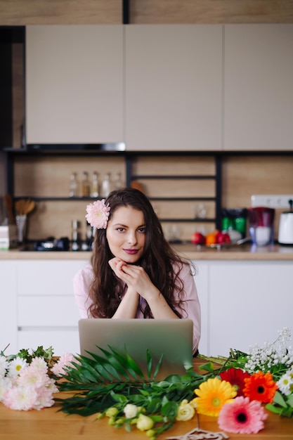 Jeune femme brune aux longs cheveux ondulés travaillant sur ordinateur portable avec des fleurs sur le bureau avec cuisine en arrière-plan Fleuriste talentueux développant des ventes en ligne se préparant pour l'atelier