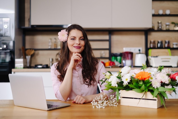 Jeune femme brune aux longs cheveux ondulés travaillant sur ordinateur portable avec des fleurs sur le bureau avec cuisine en arrière-plan Fleuriste talentueux développant des ventes en ligne se préparant pour l'atelier