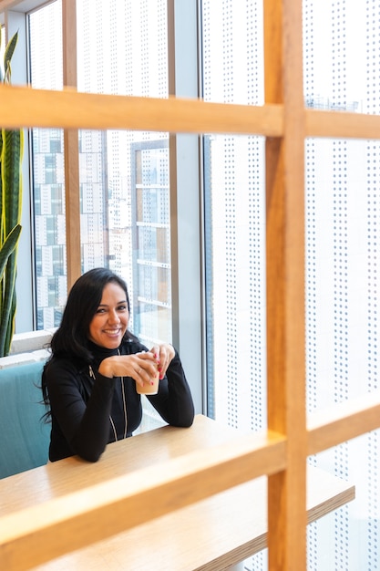 Une jeune femme brune boit du café et regarde pensivement par la fenêtre du bar de la rue tout en passant son temps seule