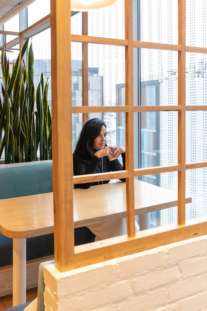 Une jeune femme brune boit du café et regarde pensivement par la fenêtre du bar de la rue tout en passant son temps seule