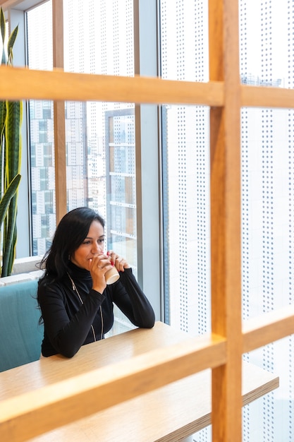 Une jeune femme brune boit du café et regarde pensivement par la fenêtre du bar de la rue tout en passant son temps seule