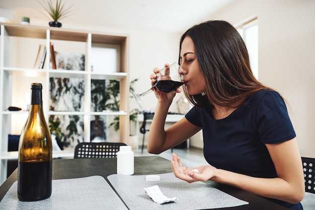 Jeune femme brune buvant un verre de vin rouge et prenant des pilules seule dans la cuisine à la maison. Concept d'alcoolisme féminin. Protestation dans le traitement de la dépendance à l'alcool.