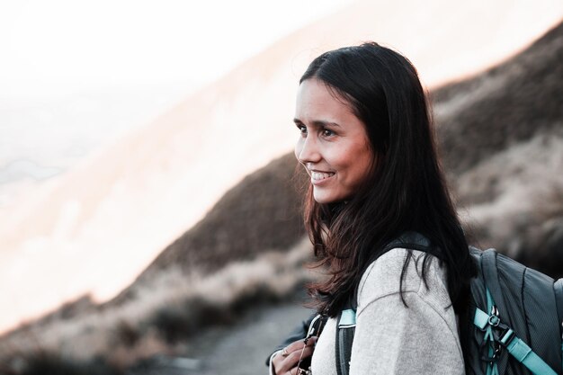 Une jeune femme brune caucasienne avec des yeux bruns, un pull gris et un sac à dos qui a l'air perdue, souriante et heureuse.