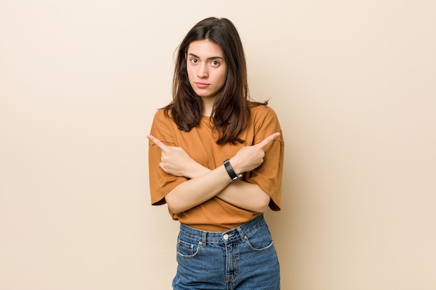 Jeune femme brune contre un mur beige pointe de côté, essaie de choisir entre deux options.