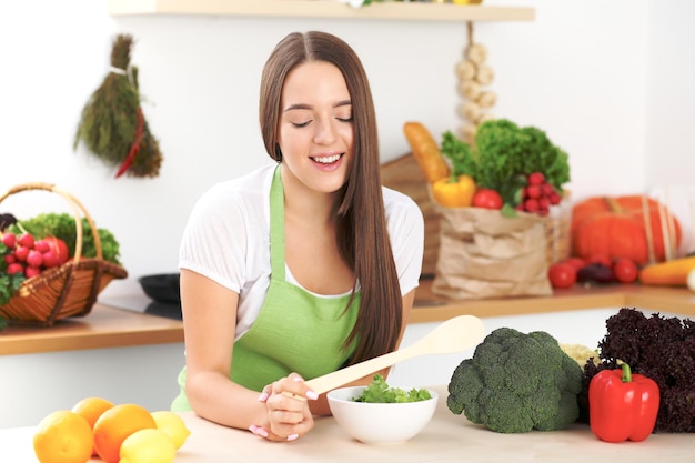 Jeune femme brune cuisine ou mange de la salade fraîche dans la cuisine. Femme au foyer tenant une cuillère en bois dans sa main droite. Concept de nourriture et de santé.