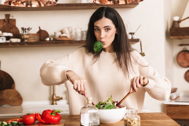 Jeune femme brune cuisiner et manger une salade verte saine avec des légumes à la maison