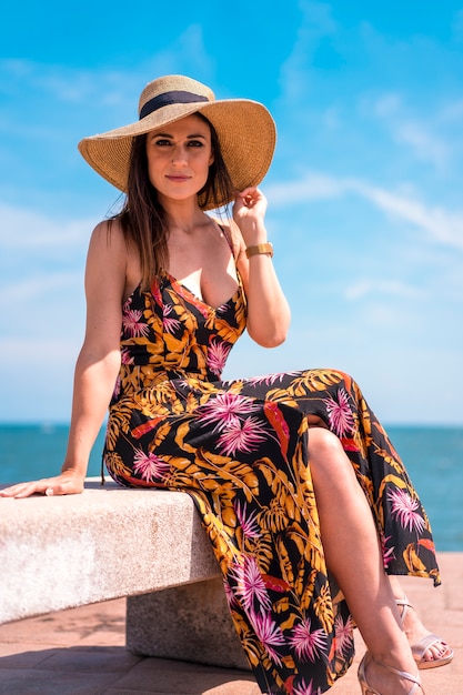 Une jeune femme brune dans une robe et un chapeau de paille au bord de la mer