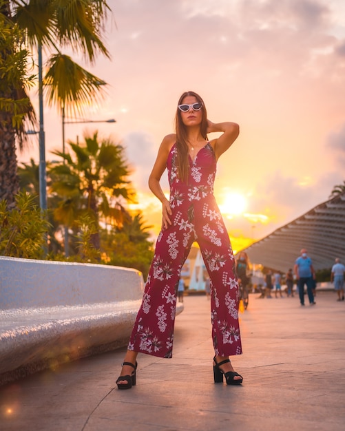 Jeune femme brune dans une robe à fleurs marron et des lunettes de soleil modernes profitant de l'été à l'heure d'or