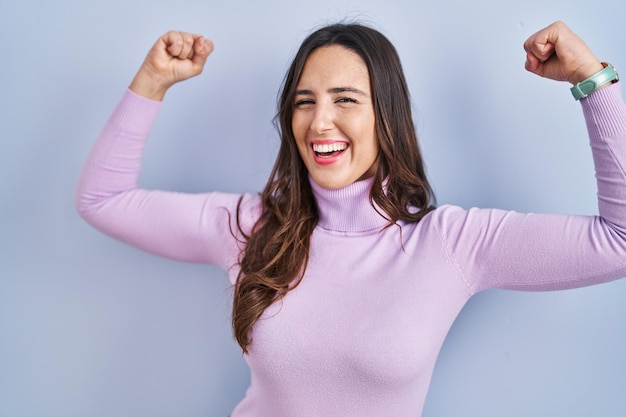 Jeune femme brune debout sur fond bleu montrant les muscles des bras souriant fier concept de remise en forme