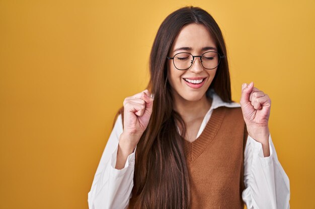 Jeune femme brune debout sur fond jaune portant des lunettes excité pour le succès avec les bras levés et les yeux fermés célébrant la victoire souriant concept gagnant