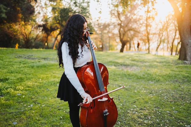 Jeune femme brune debout avec des lunettes jouant du violoncelle au coucher du soleil dans le parc, sur une herbe verte.