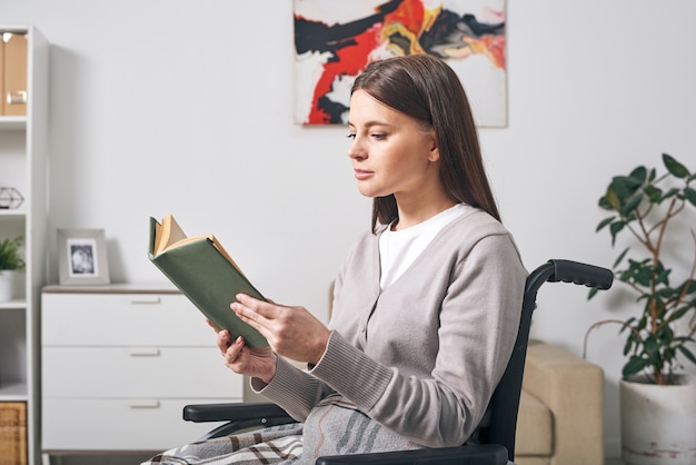 Jeune femme brune désactiver dans des vêtements décontractés assis en fauteuil roulant et livre de lecture tout en passant la journée à la maison